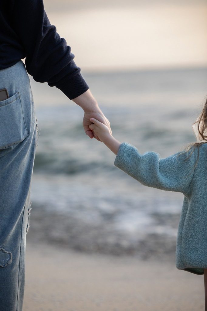 Mother holding daughters hand as they walk in the sand towards the ocean.