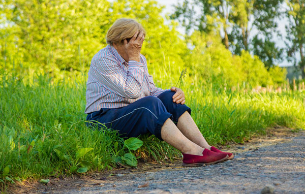 Senior sits in grass on side of road with hand on head appearing to be lost.